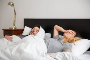 Photo by Kampus Production: https://www.pexels.com/photo/man-sleeping-beside-a-woman-7556565/ Alt: Woman frustrated with snoring partner 